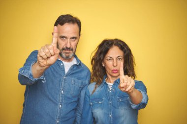 Güzel orta yaşlı çift, izole edilmiş sarı arka planda dikiliyorlar. Parmağını havaya kaldırıyorlar ve kızgın bir ifade sergiliyorlar.