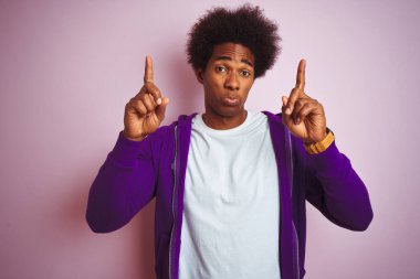 Mor kazak giyen genç Afro-Amerikalı adam pembe arka planda dikiliyor üzgün ve üzgün görünüyor, parmaklarıyla yönü gösteriyor, mutsuz ve depresif..