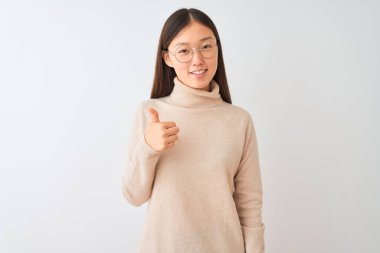 Balıkçı yaka kazak ve gözlük takan genç Çinli bir kadın izole edilmiş beyaz arka planda mutlu bir el hareketi yapıyor. Kameraya bakıp başarıyı gösteren ifade onaylanıyor.