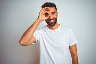İzole edilmiş beyaz arka plan üzerinde duran genç Hintli adam el hareketleriyle gülümsüyor ve mutlu yüzle parmaklarının arasından bakıyor..