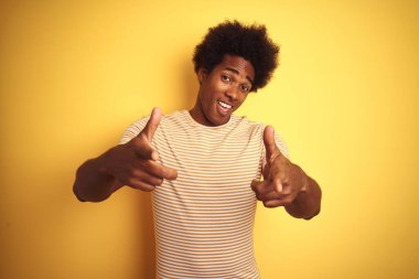 Afro saçlı, çizgili tişörtlü, izole edilmiş sarı arka plan üzerinde durup mutlu ve komik yüzlü parmakları kameraya gösteren Amerikalı bir adam. İyi enerji ve titreşimler.