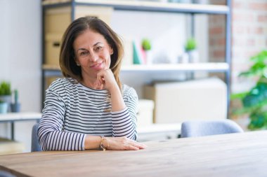 Orta yaş yaşlı kadın evde masada otururken elini çeneüzerinde sorgular, dalgın ifade. Düşünceli bir yüzle gülümsüyor. Şüphe kavramı.