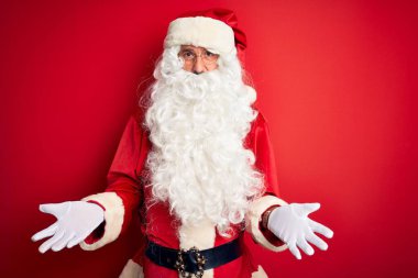 Noel Baba kostümü giymiş orta yaşlı yakışıklı adam izole edilmiş kırmızı arka planda neşeli gülümsüyor ve dostça karşılanıyor, olumlu ve kendinden emin selamlar.