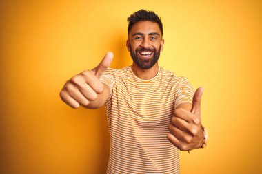 İzole edilmiş sarı arka plan üzerinde duran genç Hintli adam el hareketini onaylıyor, gülümsüyor ve başarı için mutlu. Kazanan jest.