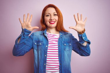Güzel kızıl saçlı kadın kot gömlek ve çizgili tişört giyerek izole pembe arka plan üzerinde ve parmaklarıyla on numarayı gösterirken kendinden emin ve mutlu bir şekilde gülümsüyor..