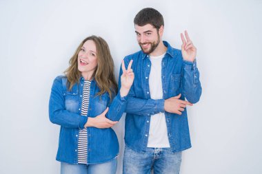 Genç güzel çift beyaz izole arka plan üzerinde birlikte ayakta mutlu yüz zafer işareti yapıyor kameraya göz kırparak gülümseyerek. İki numara.
