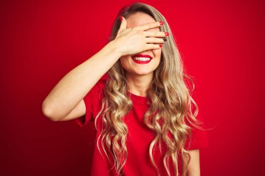 Genç ve güzel bir kadın, kırmızı izole edilmiş arka plan üzerinde dikilip gülümseyerek ve gülümseyerek gözleri kapayarak şaşkınlıktan kahkaha atıyor. Kör kavram.