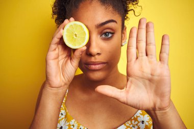 Genç Afro-Amerikalı bir kadın limon dilimini izole edilmiş sarı arka planda tutuyor. Açık elle dur işareti yapıyor. Ciddi ve kendinden emin bir ifadeyle, savunma jesti.