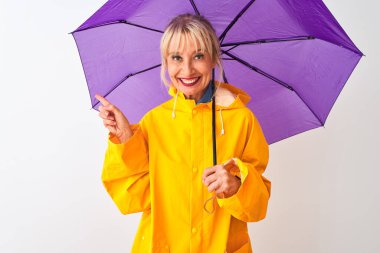 İzole edilmiş beyaz arka plan üzerinde yağmurluk ve mor şemsiye giyen orta yaşlı bir kadın elini ve parmağını kenarı işaret ederek çok mutlu olur.