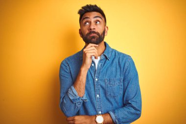 İzole edilmiş sarı arka plan üzerinde duran kot gömlek giyen genç Hintli bir adam. Elleri çenesinde soru sorarken düşünceli bir ifade takınıyor. Düşünceli bir yüzle gülümsüyor. Şüphe kavramı.