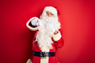 Noel Baba kostümü giymiş orta yaşlı yakışıklı bir adam izole edilmiş kırmızı arka planda durmuş sana gülüyor, eli ağzında kamerayı işaret ediyor, yüz ifadesi utanç verici.