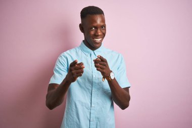 İzole edilmiş pembe arka planda dikilip mutlu ve komik yüzlü parmakları kameraya gösteren mavi gömlekli Afro-Amerikalı bir adam. İyi enerji ve titreşimler.