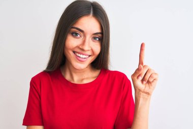 İzole edilmiş beyaz arka planda kırmızı tişört giyen genç ve güzel bir kadın elini ve parmağını kenarı göstererek mutlu bir şekilde dikiliyor.