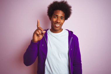 Mor kazak giyen genç Afro-Amerikalı adam pembe arka planda duruyor. Bir numarayı işaret ediyor ve gülümsüyor. Kendinden emin ve mutlu..