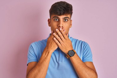 İzole edilmiş pembe arka planda duran mavi polo giyen genç Hintli adam yanlışlıkla ağızlarını kapatarak şok geçirdi. Gizli konsept.