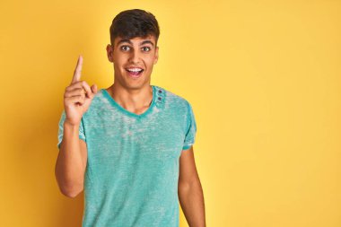 Yeşil t-shirt giyen genç Hintli adam izole edilmiş sarı arka planda duruyor ve başarılı bir fikri işaret ediyor. Heyecanlı ve mutlu. Bir numara..