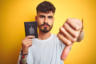 Kanada pasaportu takan genç adam izole edilmiş sarı arka planda kızgın yüzlü, olumsuz işaretli baş parmakları aşağıda, reddedilme konsepti var.
