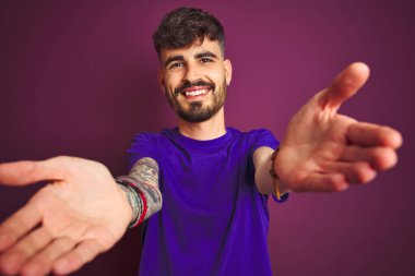 İzole edilmiş mor arka planda duran tişörtlü genç adam sarılmak için kollarını açmış gülümseyen kameraya bakıyor. Mutluluğu kucaklayan neşeli bir ifade..