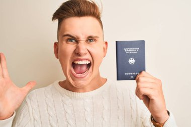 Genç yakışıklı adam Almanya pasaportunu izole edilmiş beyaz arka planda tutuyor çok mutlu ve heyecanlı, kazanan yüz ifadesi zaferi büyük bir gülümsemeyle kutluyor ve ellerini kaldırıyor.
