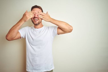 İzole edilmiş arka plan üzerine gündelik beyaz tişört giyen genç yakışıklı adam neşeli ve komik gülümseyen elleriyle gözlerini kapatıyor. Kör kavram.