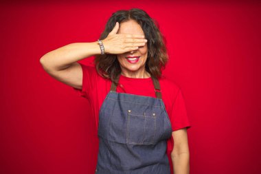 Kırmızı izole edilmiş arka planda önlük giymiş orta yaşlı bir kadın. Gülümsüyor ve yüzünde şaşkınlıktan gözleri kapayarak gülüyor. Kör kavram.