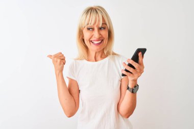 Akıllı telefon kullanan orta yaşlı bir kadın izole edilmiş beyaz arka planda duruyor ve gülümseyen bir gülümsemeyle baş parmağıyla işaret ediyor.