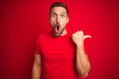 Kırmızı izole edilmiş arka plan üzerine gündelik tişört giyen genç yakışıklı bir adam. El parmağı yana dönük, ağzı açık, şaşkın bir ifade..