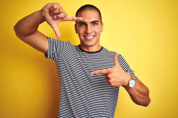 黄色の孤立した背景にストライプのTシャツを着た若いハンサムな男は 幸せな顔をした手と指でフレームを作り笑顔 創造性と写真の概念 — ストック写真