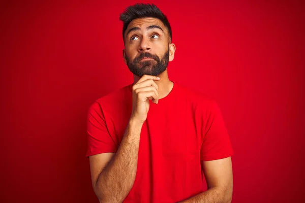 Young handsome indian man wearing t-shirt over isolated red background with hand on chin thinking about question, pensive expression. Smiling with thoughtful face. Doubt concept.