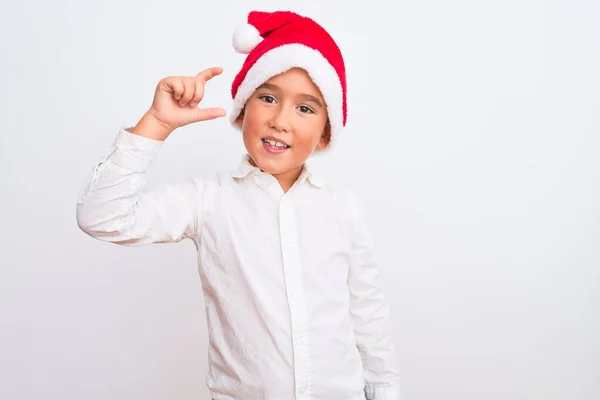 孤立した白い背景の笑顔の上に立つクリスマスサンタの帽子を身に着けている美しい少年と指を探しているとカメラと小さなサイズの看板を行う手で自信を持ってジェスチャー 概念の測定 — ストック写真