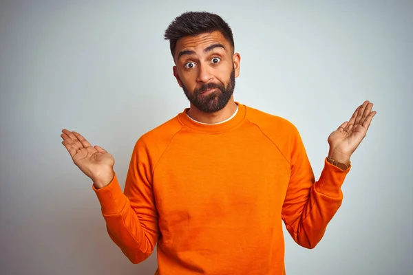 孤立した白い背景にオレンジ色のセーターを着た若いインド人男性は 腕や手を上げて混乱した表情をしていました 疑いの概念 — ストック写真
