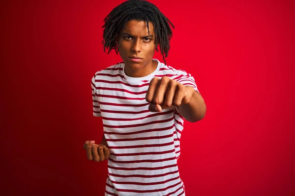 若いですアフロ男ですストライプTシャツを身に着けています独立した赤い背景の上に立って戦いに拳をパンチ 積極的かつ怒りの攻撃 脅威と暴力 — ストック写真