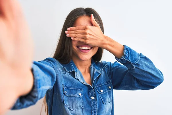 Beautiful woman wearing denim shirt make selfie by camera over isolated white background smiling and laughing with hand on face covering eyes for surprise. Blind concept.