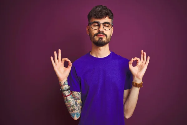 穿着T恤衫和眼镜站在孤立的紫色背景上的年轻男子放松 闭上双眼微笑 用手指做冥想动作 瑜伽概念 — 图库照片