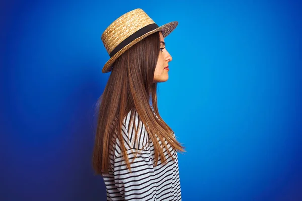 Young beautiful woman wearing navy striped t-shirt and hat over isolated blue background looking to side, relax profile pose with natural face with confident smile.