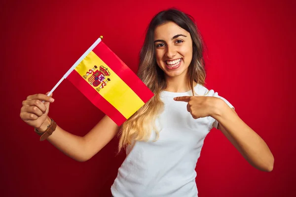 Young beautiful woman holding spanish flag over red isolated background with surprise face pointing finger to himself