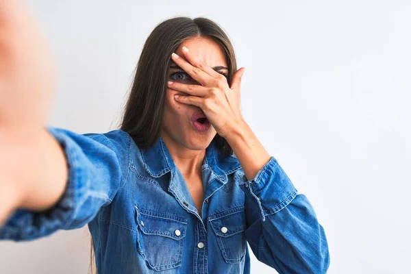 Beautiful woman wearing denim shirt make selfie by camera over isolated white background peeking in shock covering face and eyes with hand, looking through fingers with embarrassed expression.