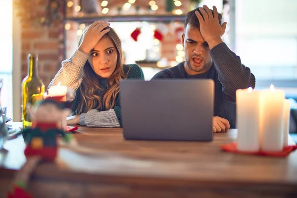 Young beautiful couple sitting using laptop around christmas decoration at home surprised with hand on head for mistake, remember error. Forgot, bad memory concept.