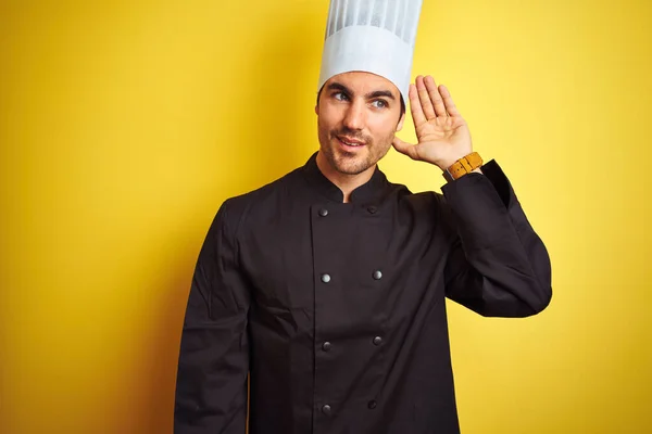 年轻厨师 身穿制服 头戴帽子 站在孤立的黄色背景上 面带微笑 两手相对地听着谣言或流言蜚语 失聪的概念 — 图库照片