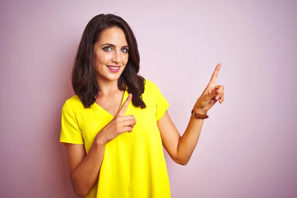 年轻美丽的女人穿着黄色的T恤 站在粉色孤立的背景上微笑着 看着摄像机 用双手和手指指向旁边 — 图库照片