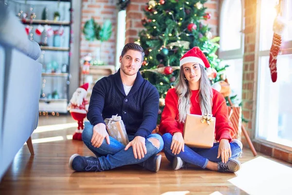 一对年轻的夫妇头戴圣爪帽 坐在家里圣诞树下的地板上 脸上带着严肃的表情 简单而自然地看着相机 — 图库照片