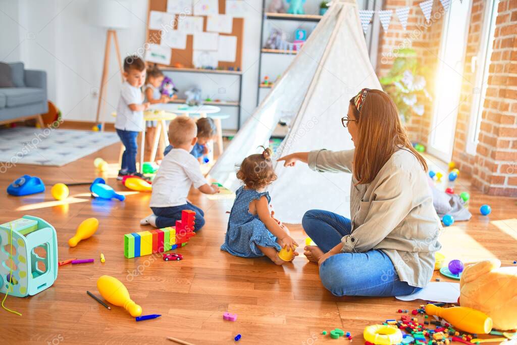 Beautiful teacher and group of toddlers playing around lots of toys at kindergarten