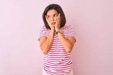 İzole edilmiş pembe arka plan üzerinde duran çizgili tişört giyen genç ve güzel bir kadın. Yüzü, depresyonu ve üzüntüyü örten yorgun eller, sorun yüzünden üzgün ve tedirgin.