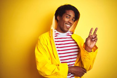 Siyah saçlı, yağmurluk giyen, izole edilmiş sarı arka planda duran, kameraya gülümseyen ve zafer işareti yapan Afrikalı bir adam. İki numara..