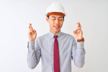 Kravat ve kask takan Çinli mimar izole edilmiş beyaz arka planda durup umut ve gözlerin kapalı bir şekilde gülümseyerek işaret ediyor. Şans ve batıl inanç..