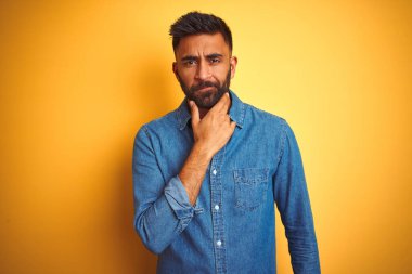 İzole edilmiş sarı arka planda duran kot gömlek giyen genç Hintli adam kameraya kollarını kavuşturup gülümseyerek ve çenesini kaldırarak bakıyor. Olumlu düşünüyorum..