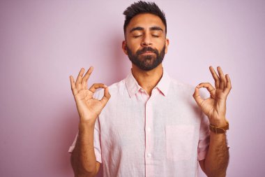 Izole pembe arka plan üzerinde duran rahat gömlek giyen genç hintli adam rahatlayın ve parmakları ile meditasyon jest yapıyor kapalı gözleri ile gülümseyerek. Yoga konsepti.