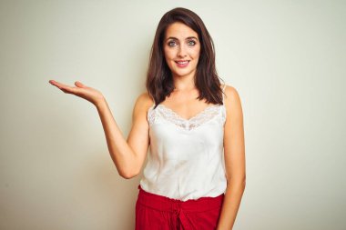 Tişört giyen genç ve güzel bir kadın izole edilmiş beyaz arka planda dikiliyor. Gülümsüyor, mutlu bir şekilde sunuyor ve avuç içi kameraya bakıyor..