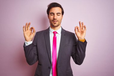 Genç yakışıklı iş adamı takım elbise ve kravat takıyor pembe arka planda dikiliyor rahatlıyor ve parmaklarıyla meditasyon yaparken gözleri kapalı gülümsüyor. Yoga konsepti.