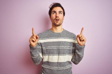 İzole edilmiş pembe arka plan üzerinde duran sıradan çizgili süveter giyen genç yakışıklı adam şaşırmış ve şaşırmış bir şekilde yukarı bakıyor ve parmaklarıyla işaret ediyor ve kollarını kaldırıyor..
